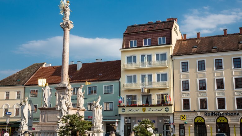Ein Platz mit einer S&auml;ule und Statuen, umgeben von Geb&auml;uden, darunter das Hotel Zentral.