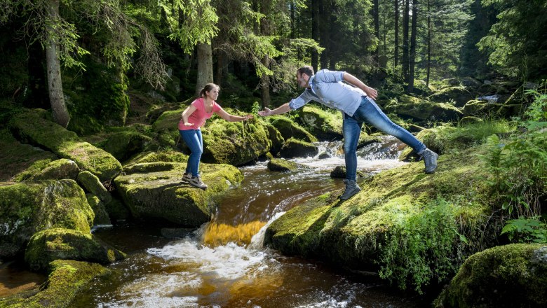 Zwei Personen stehen auf moosbedeckten Steinen &uuml;ber einem Bach in einem Wald und reichen sich die H&auml;nde.