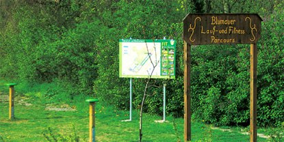 Holzschild mit der Aufschrift 'Blumauer Lauf- und Fitness Parcours' vor einer Infotafel im Gr&uuml;nen.