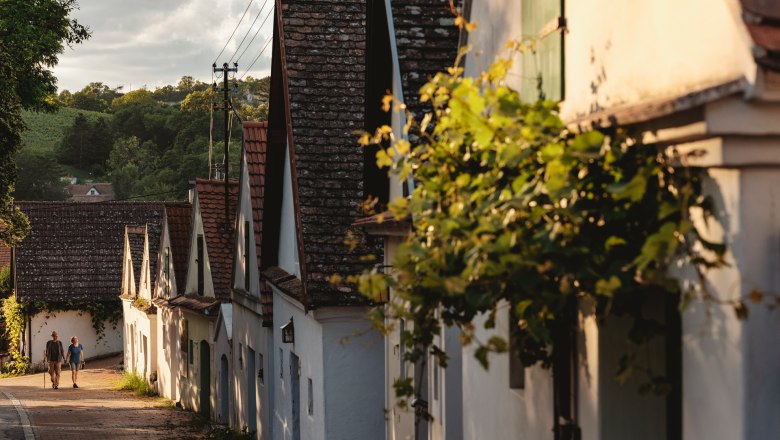Eine malerische Kellergasse in Falkenstein mit traditionellen Weinkellern und zwei Personen, die entlang der Stra&szlig;e spazieren.