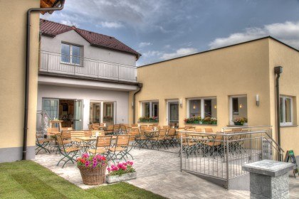 Ein gem&uuml;tlicher Au&szlig;enbereich mit Holztischen und -st&uuml;hlen vor einem gelben Geb&auml;ude.