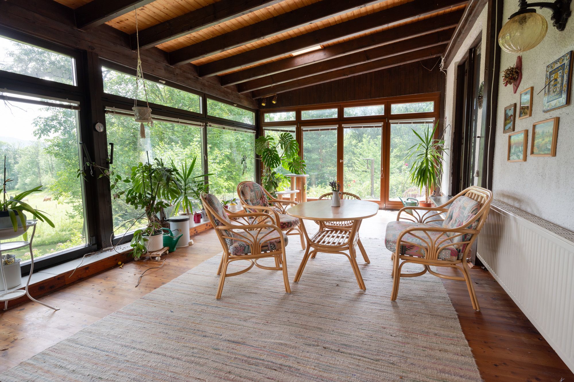 Ein Wintergarten mit Holzboden, Rattanmöbeln und vielen Pflanzen. Große Fenster bieten Blick auf grüne Natur.