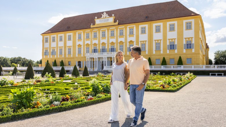 Paar spaziert neben einem Blumenbeet und im Hintergrund sieht man Schloss Hof
