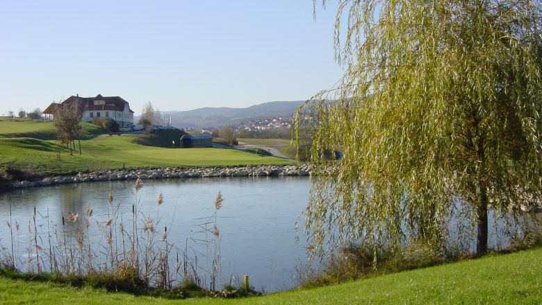 Landschaft mit Golfplatz, Teich und Gebäude im Hintergrund.