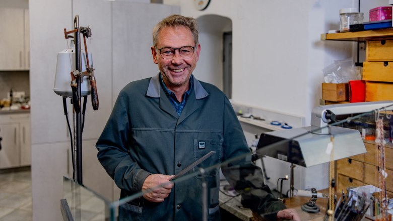 Ein Mann in einer Werkstatt mit Brille und Arbeitskittel, lächelnd, umgeben von Werkzeugen und Schmuck.