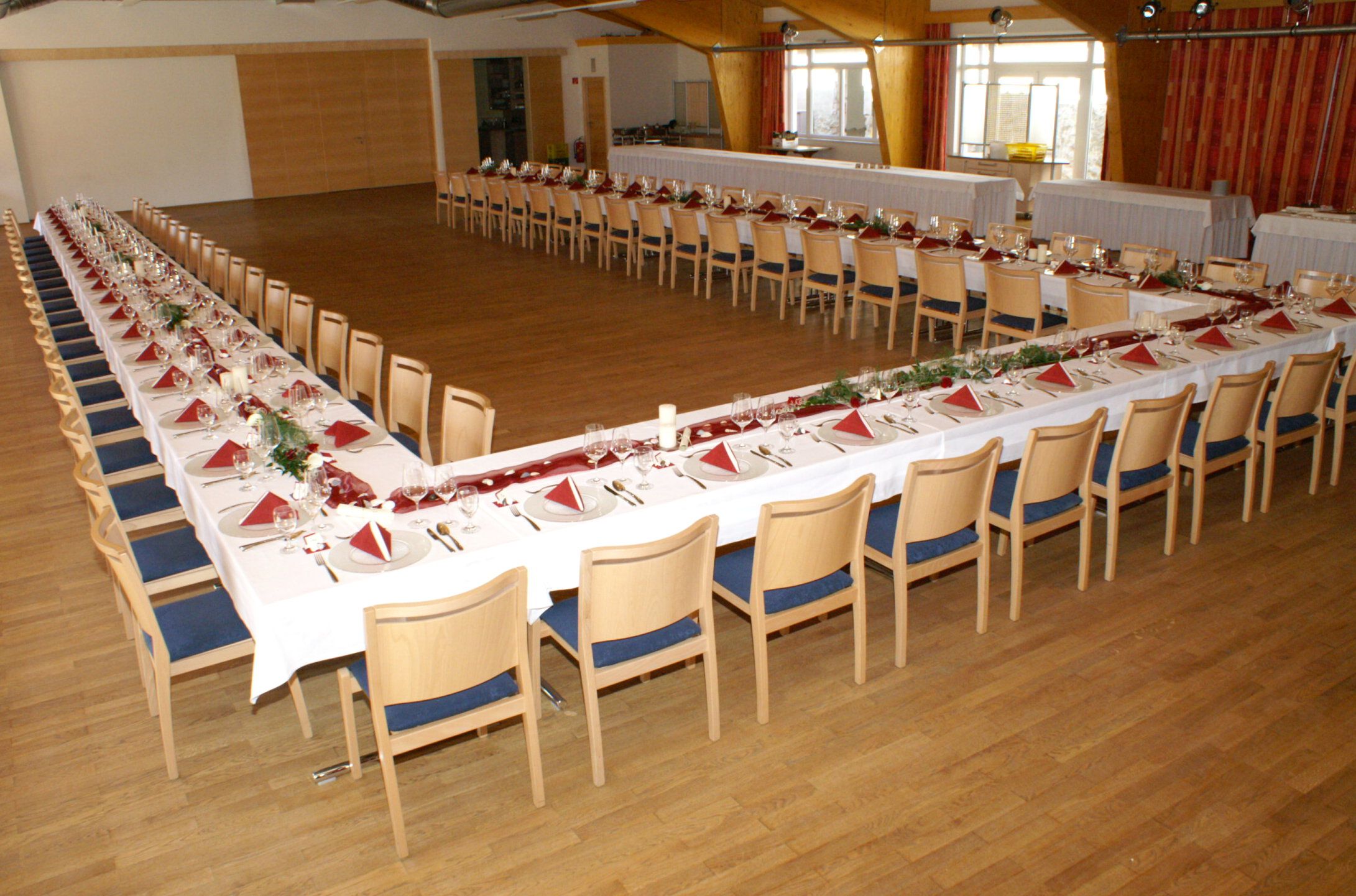 Ein großer Festsaal mit U-förmig gedeckten Tischen, dekoriert mit roten Servietten und Blumen.