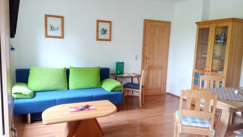 Wohnzimmer mit blauer Couch, Holztisch und St&uuml;hlen, Schrank und Bildern an der Wand.