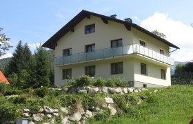 Ein zweistöckiges Haus mit Balkon in einer grünen, hügeligen Landschaft.