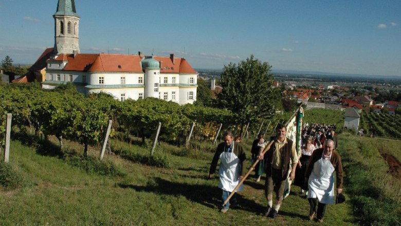 Menschen in traditioneller Kleidung gehen durch Weinberge in Gumpoldskirchen, im Hintergrund eine Kirche und Gebäude.