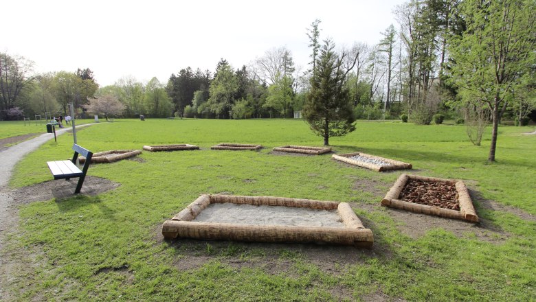 Ein Stadtpark mit einem Barfu&szlig;pfad aus verschiedenen Materialien und einer Parkbank.