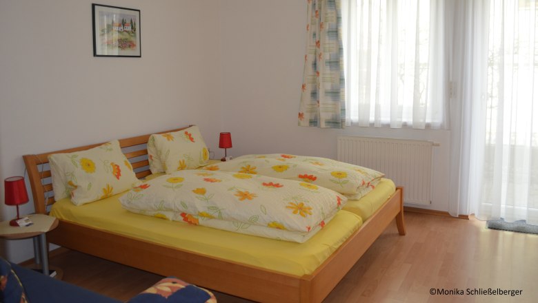 Doppelzimmer, © Monika Schließelberger Ein gemütliches Doppelzimmer mit einem Holzbett, bunter Bettwäsche und zwei roten Nachttischlampen. Ein Fenster mit Vorhängen ist im Hintergrund.