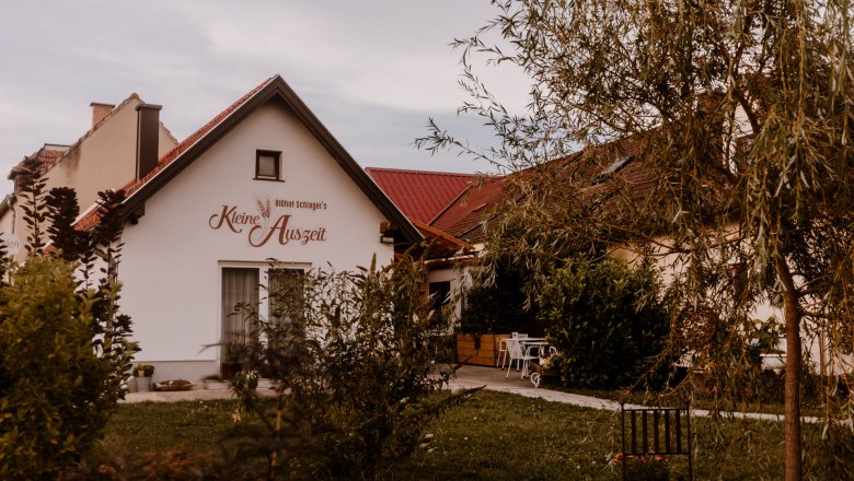 Ein kleines Geb&auml;ude mit der Aufschrift 'Biohof Schlager's Kleine Auszeit', umgeben von B&auml;umen und Gartenm&ouml;beln.