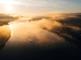 Sanfte Nebelschwaden umh&uuml;llen den ruhigen Fluss, w&auml;hrend die ersten Sonnenstrahlen den Tag erhellen. Die malerische Landschaft l&auml;dt Wanderer ein, die Sch&ouml;nheit der Natur zu entdecken und die frische Morgenluft zu genie&szlig;en.