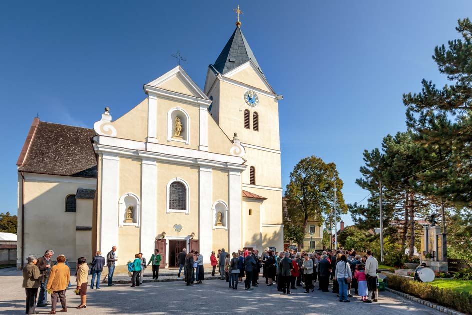 Menschen versammeln sich vor einer Kirche mit Turm und Uhr bei sonnigem Wetter.