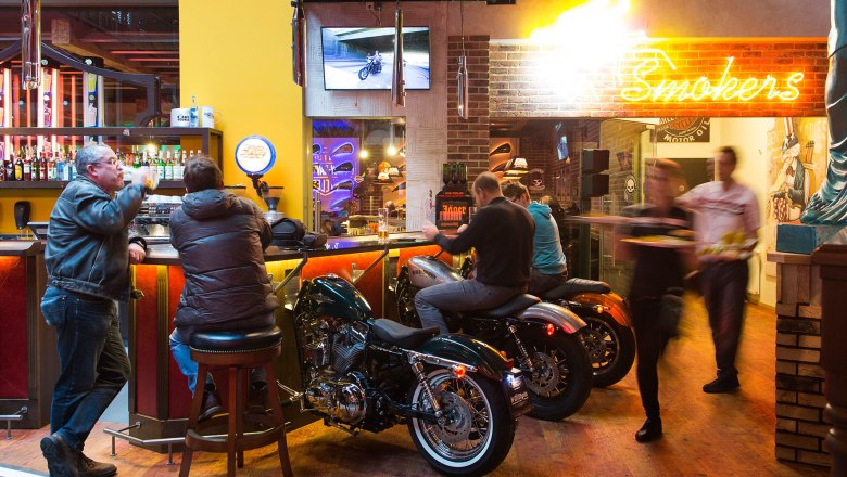 Bar mit Motorrädern als Sitzgelegenheiten, Neonlicht und Gästen.