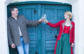 Ein Mann und eine Frau stoßen mit Weingläsern an, vor einer blauen Holztür.