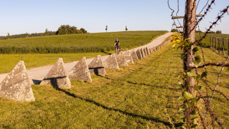 Ein Radfahrer auf einem Weg entlang alter Betonbarrieren und Stacheldraht, symbolisch f&uuml;r den Eisernen Vorhang.