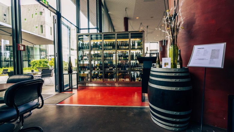 Innenansicht eines modernen Weinkellers mit Glasvitrine voller Weinflaschen und einem Weinfass als Dekoration.