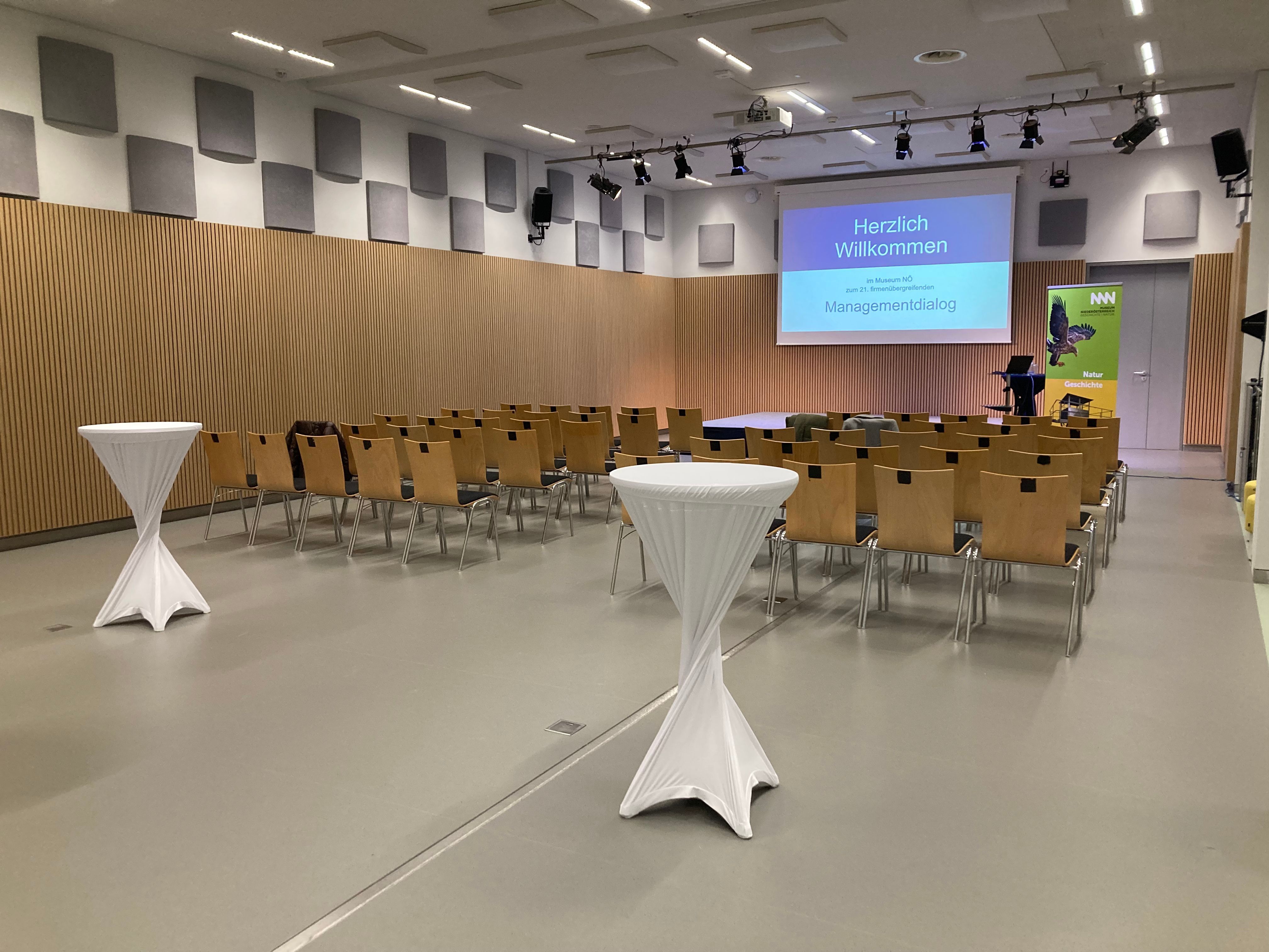 Konferenzraum mit Stuhlreihen, Stehtischen und einer Leinwand mit der Aufschrift 'Herzlich Willkommen'.