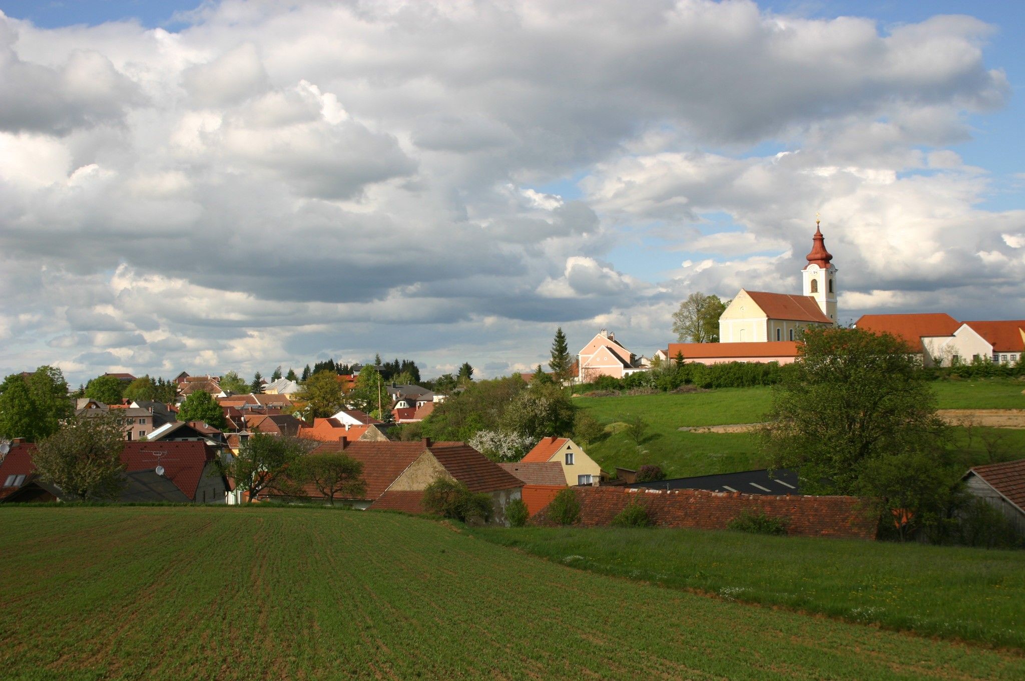 Landschaft mit Dorf und Kirche unter bewölktem Himmel.
