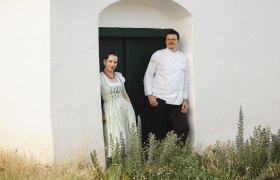 Zwei Personen stehen in einem T&uuml;rrahmen vor einem wei&szlig;en Geb&auml;ude.