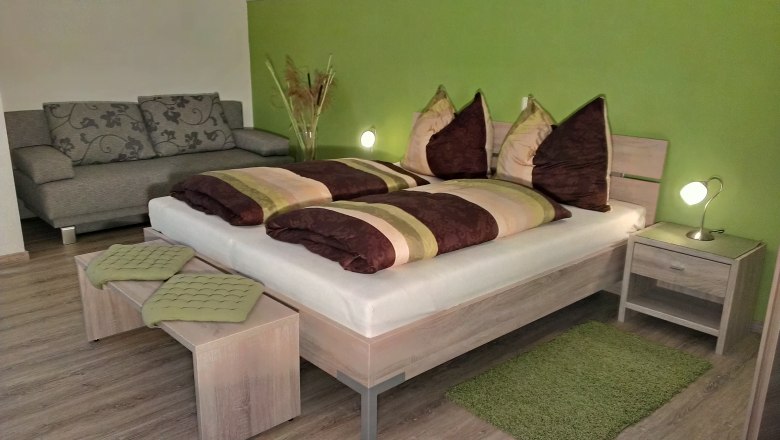 Gem&uuml;tliches Schlafzimmer mit gr&uuml;n gestrichener Wand, Doppelbett, Nachttischlampen und grauem Sofa.