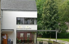 Modernes Geb&auml;ude mit der Aufschrift 'Gasthaus Burgkeller', umgeben von B&auml;umen.