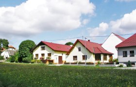 Ein Biohof mit mehreren gelben Geb&auml;uden und roten D&auml;chern, umgeben von gr&uuml;nen Wiesen und B&auml;umen unter einem blauen Himmel mit Wolken.