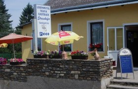 Außenansicht eines gelben Gebäudes mit Schildern für Lebensmittel, Trafik und Gasthaus. Vor dem Haus stehen Sonnenschirme und Blumen.