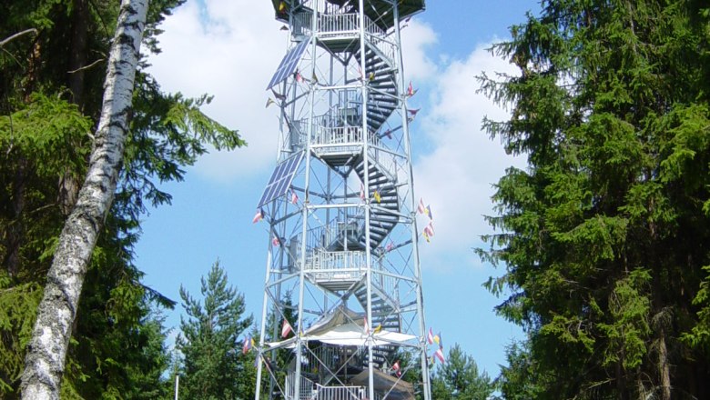 Ein hoher Aussichtsturm aus Metall mit Holzverkleidung, umgeben von B&auml;umen, unter blauem Himmel.