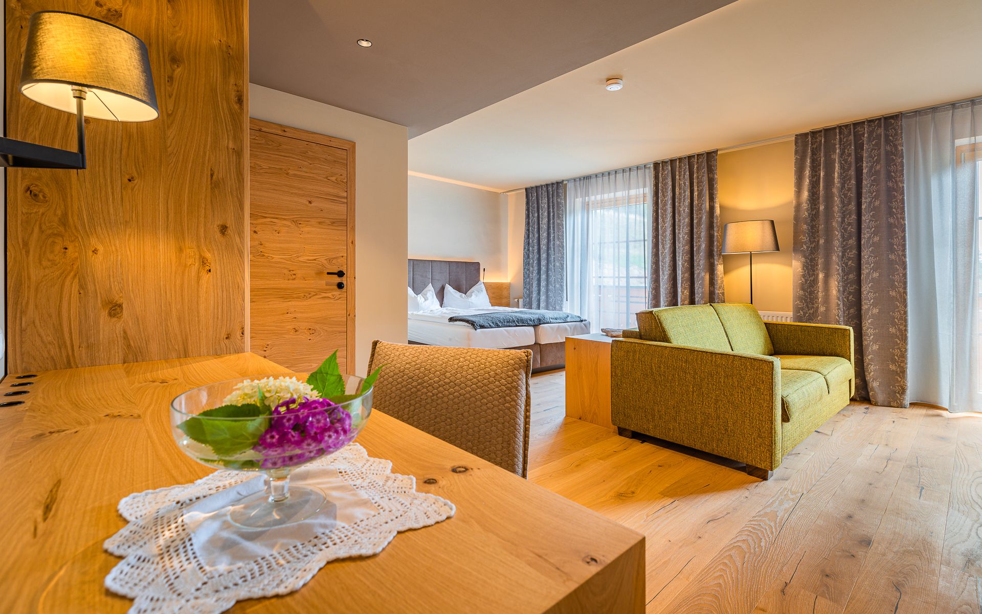 Modernes Hotelzimmer mit Holzinterieur, Bett, grünem Sofa, Tischlampe und Tisch mit Blumenschale.