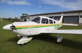 Ein kleines Flugzeug, Piper Cherokee Archer II, steht auf einer gr&uuml;nen Wiese vor einem Hangar und einem Kontrollturm.