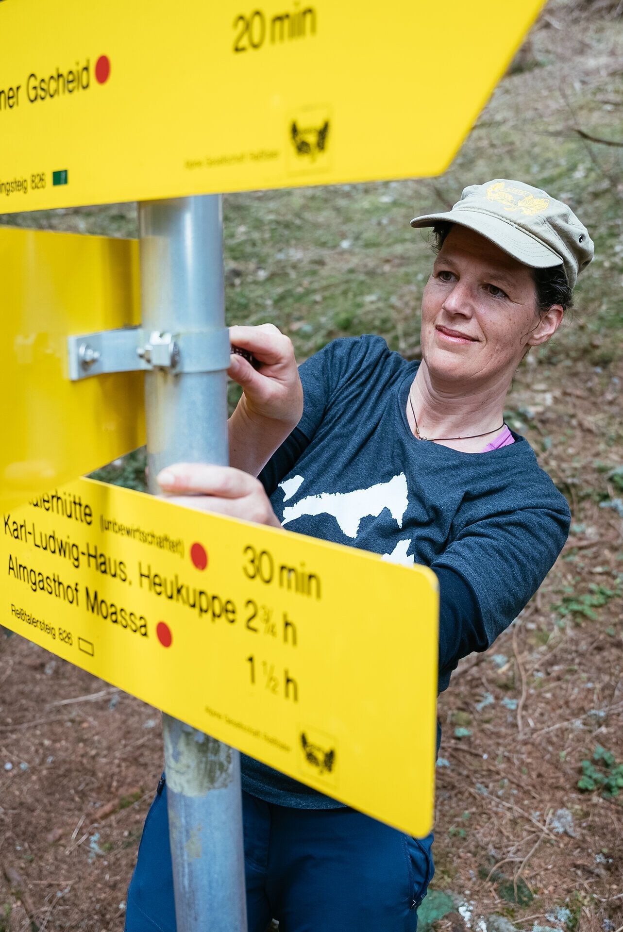 Inmitten der beeindruckenden Berglandschaft wird die Wanderin aktiv, um die Wegweiser für die nächsten Abenteurer zu justieren. Die frische Luft und die umgebende Natur laden dazu ein, die Schönheit der Alpen zu genießen und neue Wege zu erkunden.