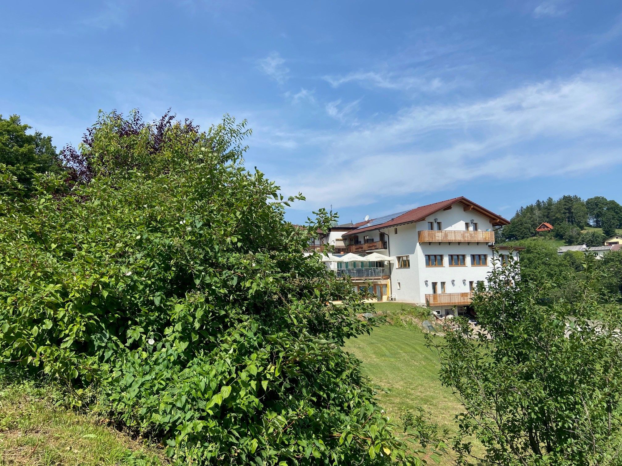 Ein weißes Haus mit rotem Dach, umgeben von grünen Bäumen und Wiesen, unter blauem Himmel.