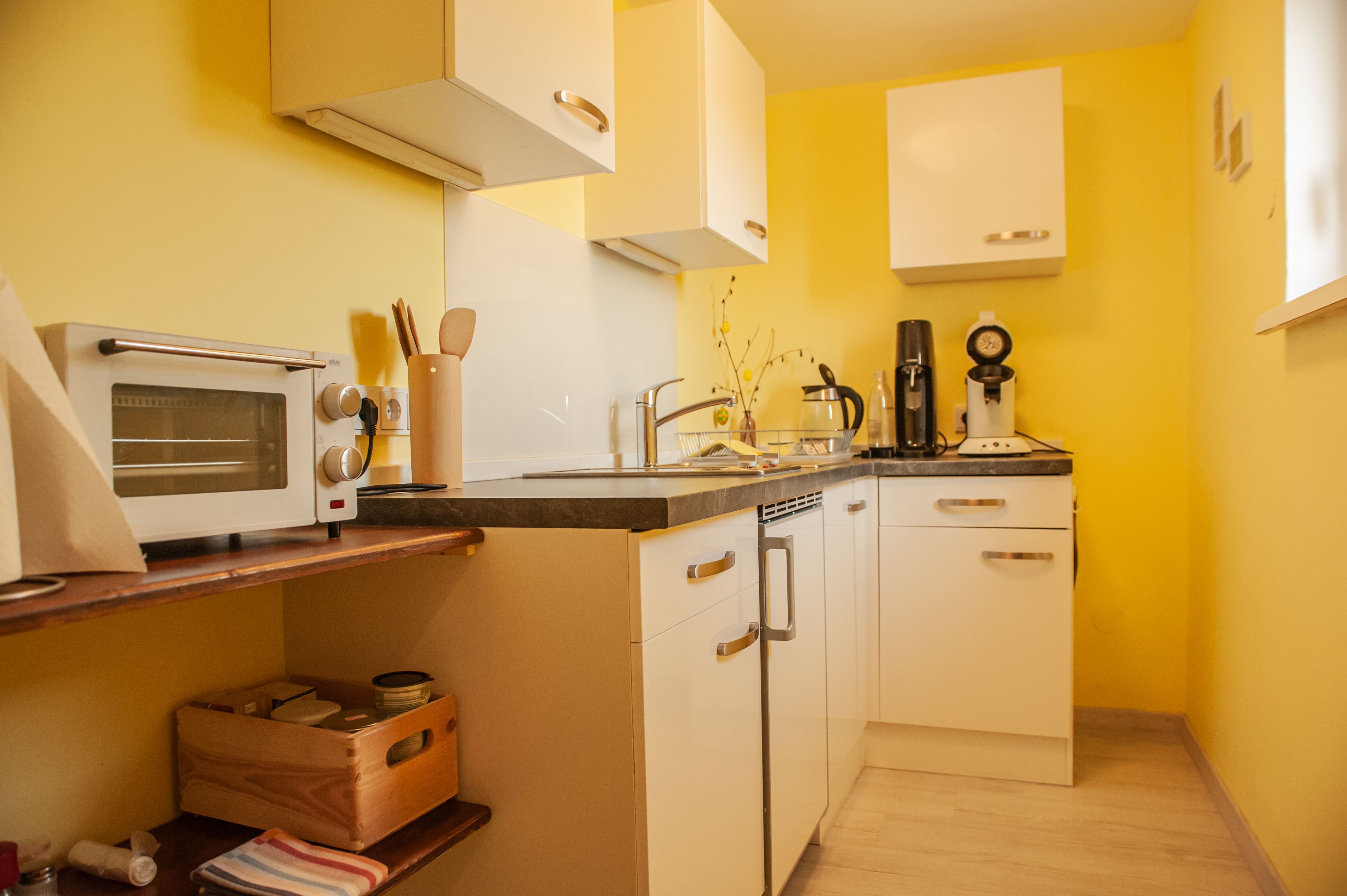 Kleine Küche mit gelben Wänden, weißen Schränken und Geräten wie Toaster und Kaffeemaschine.