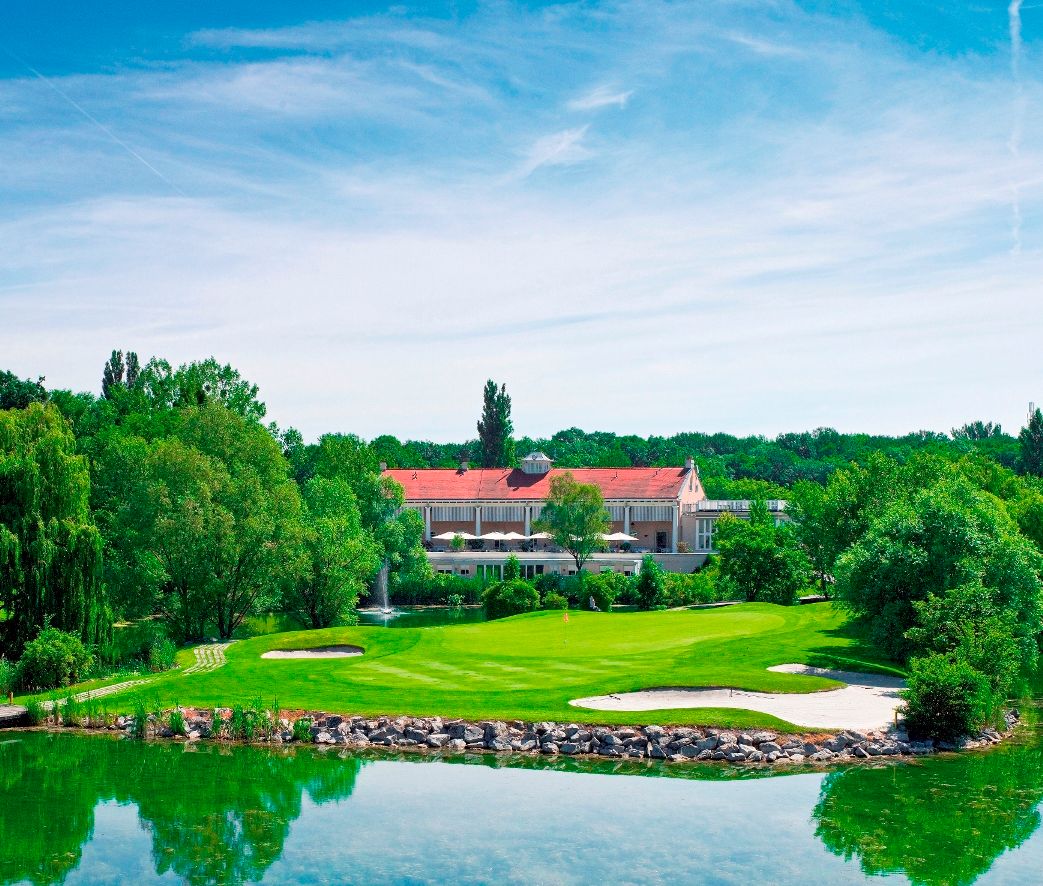 Golfplatz mit Clubhaus im Hintergrund, umgeben von Bäumen und einem Teich.
