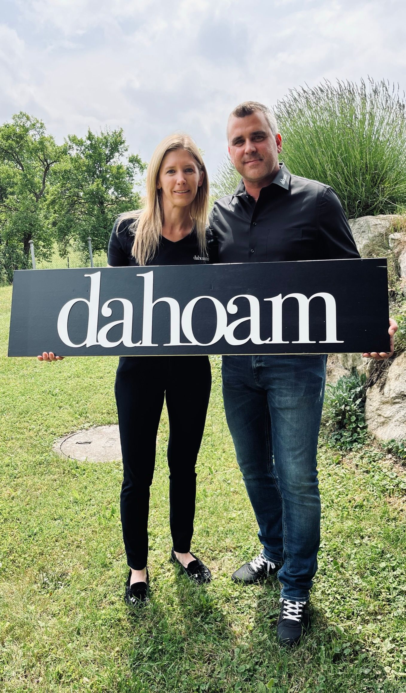 Zwei Personen halten ein Schild mit der Aufschrift 'dahoam' im Freien.