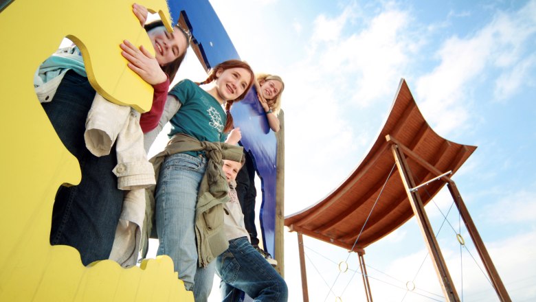 Kinder spielen auf einem bunten Kletterger&uuml;st mit einem Holzdach im Hintergrund.