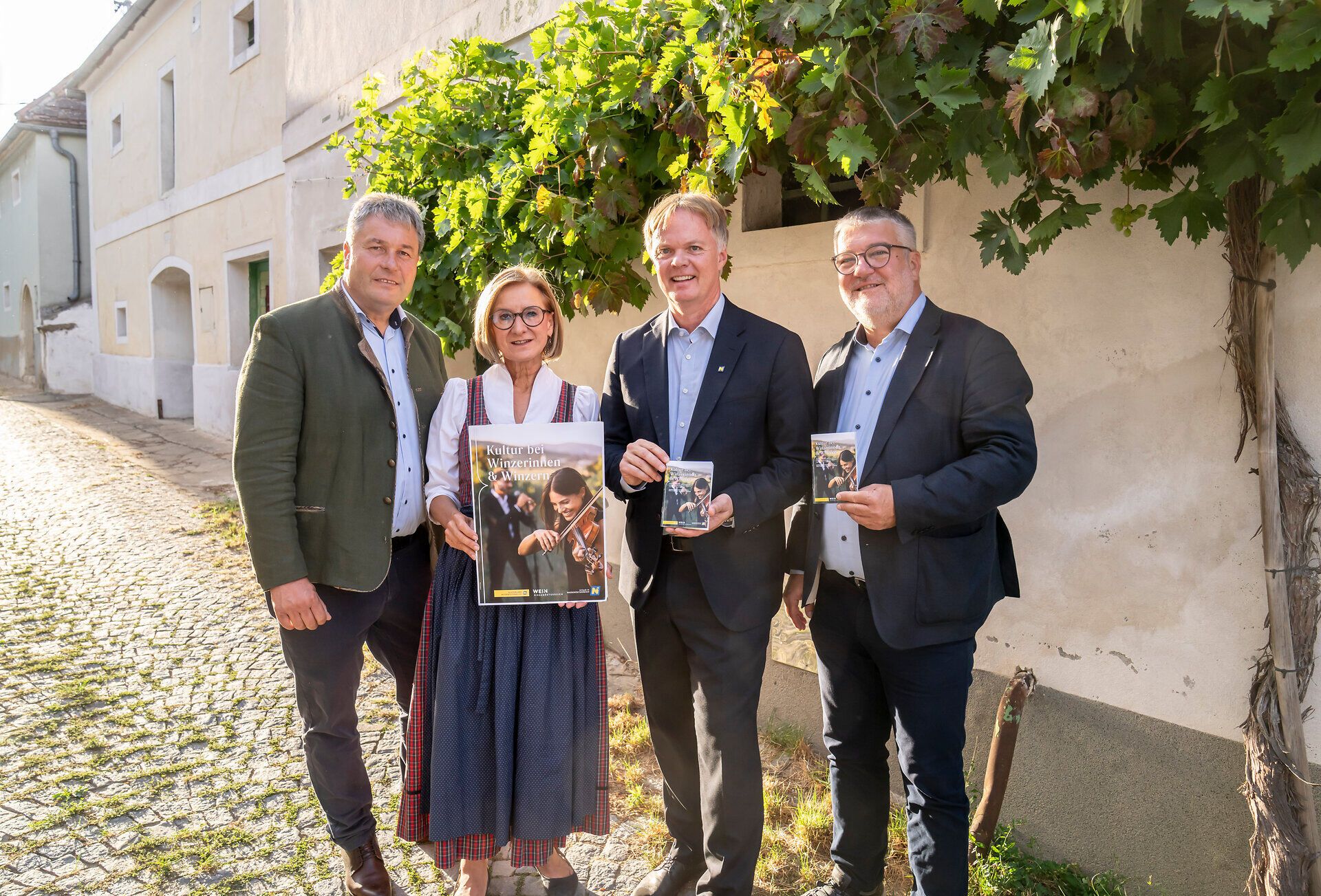 Vier Personen stehen in einer sonnigen Kellergasse unter Weinreben und präsentieren Folder bzw. Plakat zur Veranstaltungsreihe „Kultur bei Winzerinnen & Winzern“.