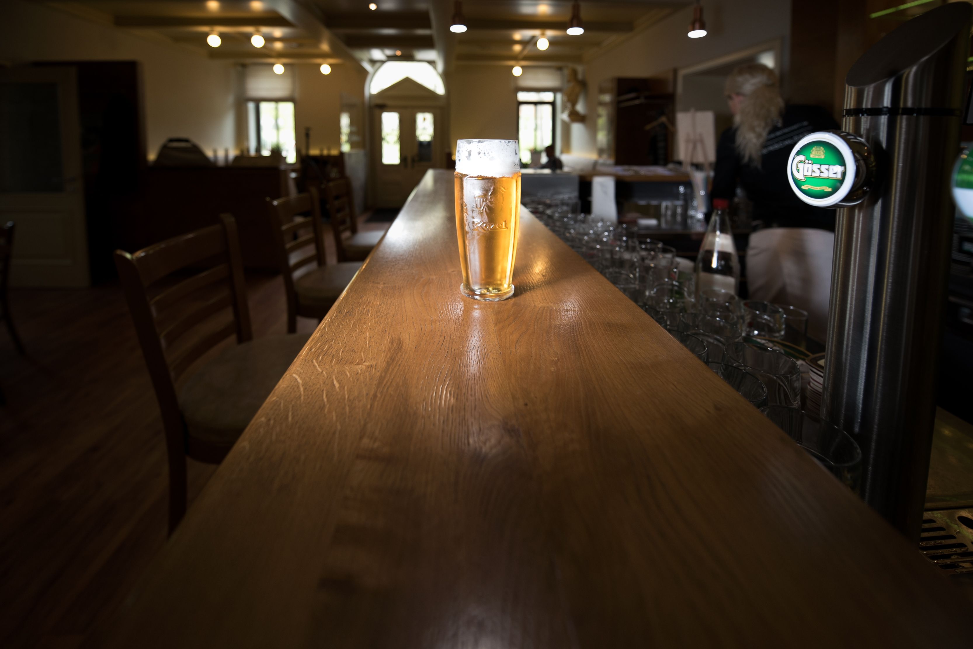 Ein Glas Bier steht auf einer langen Holztheke in einer Bar mit leeren Stühlen und einer Person im Hintergrund.