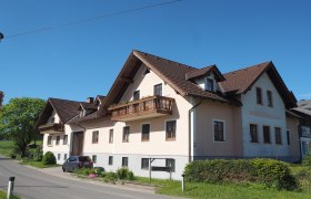 Ein großes Bauernhaus mit braunem Dach und Balkonen an einer Landstraße bei klarem Himmel.