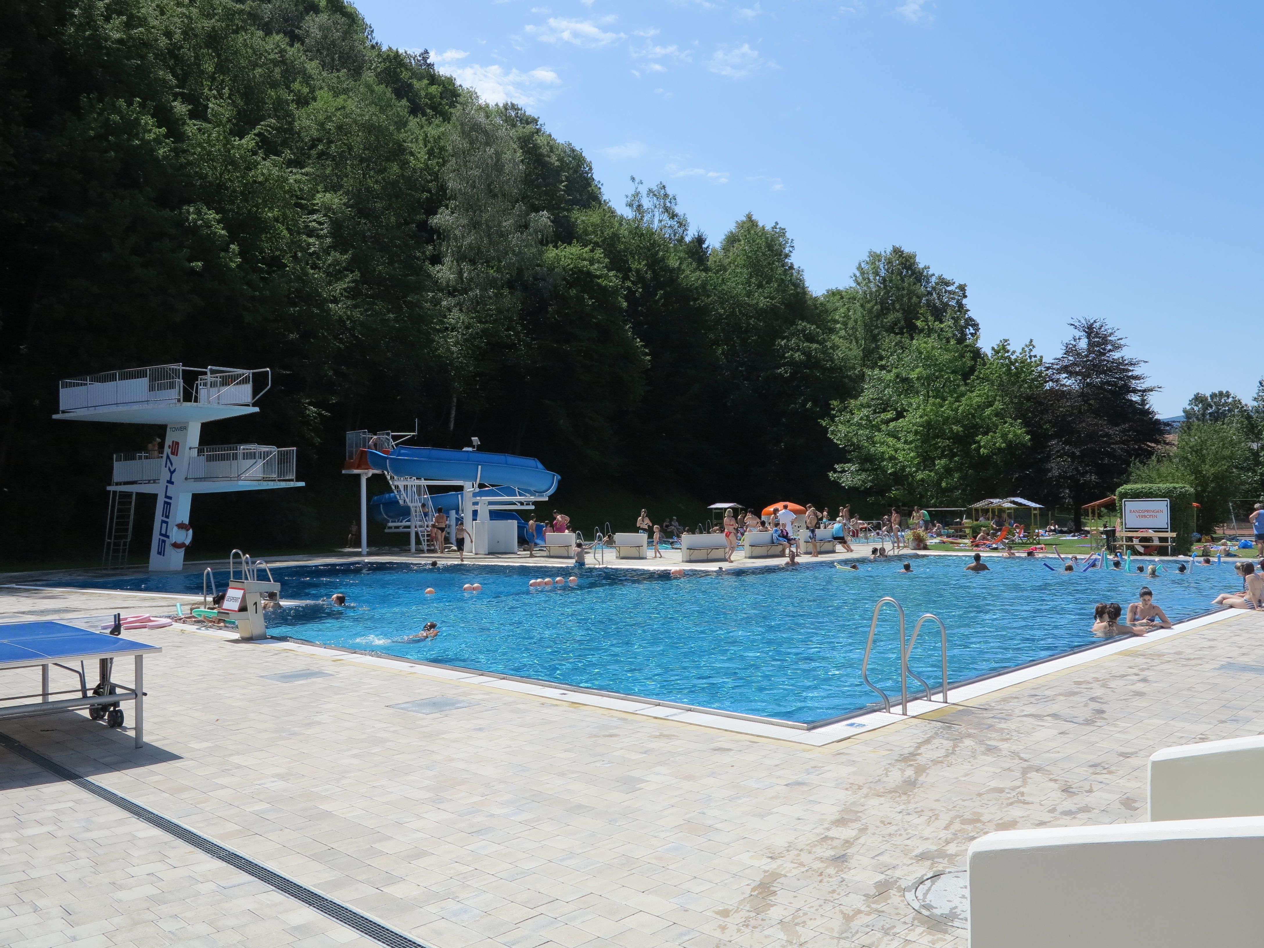 Ein Freibad mit Sprungturm, Rutsche und vielen Badegästen an einem sonnigen Tag.