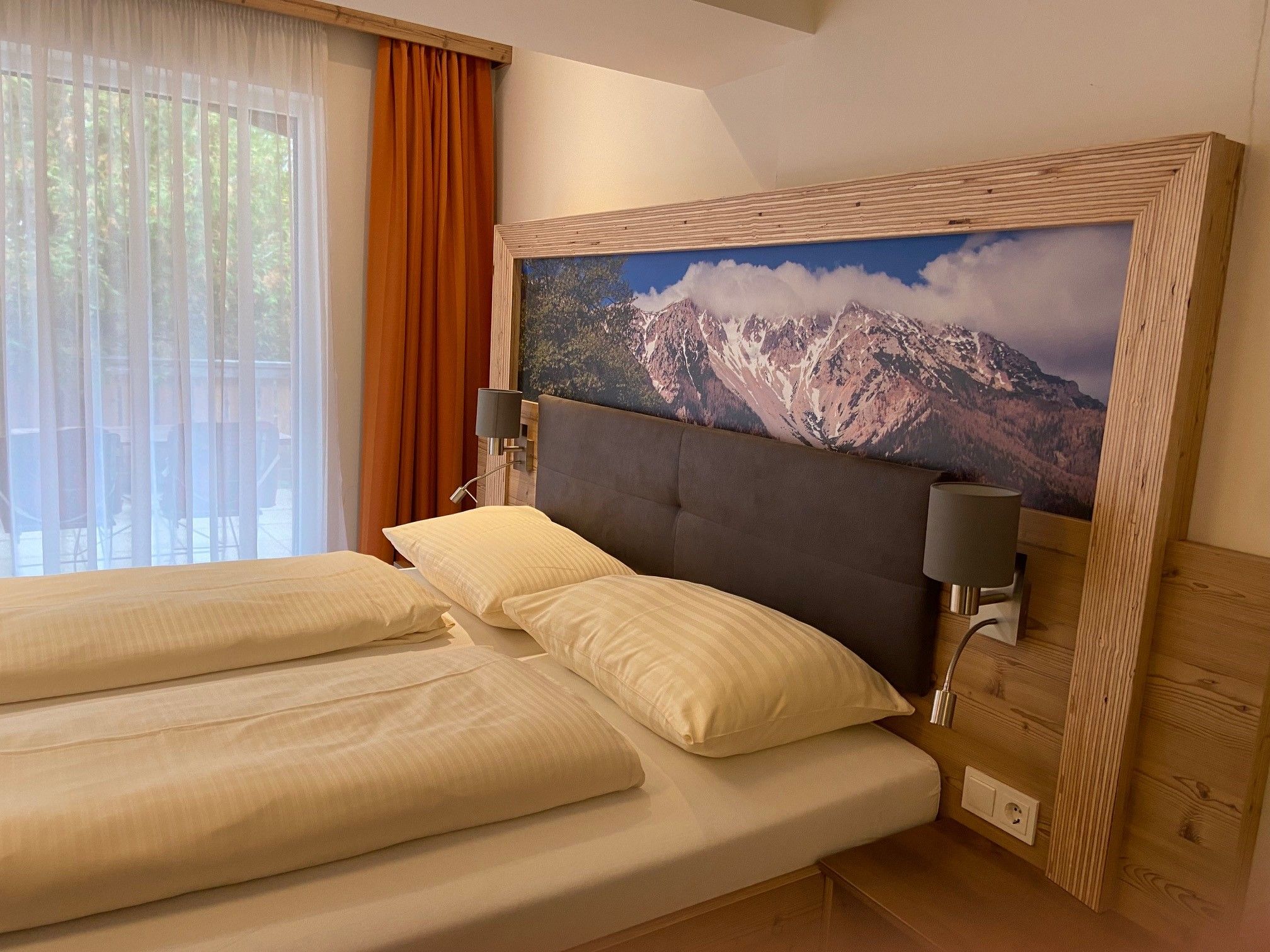 Ein gemütliches Doppelzimmer mit einem großen Bett, Holzmöbeln und einem Bild von Bergen über dem Kopfteil. Ein Fenster mit Vorhängen ist zu sehen.