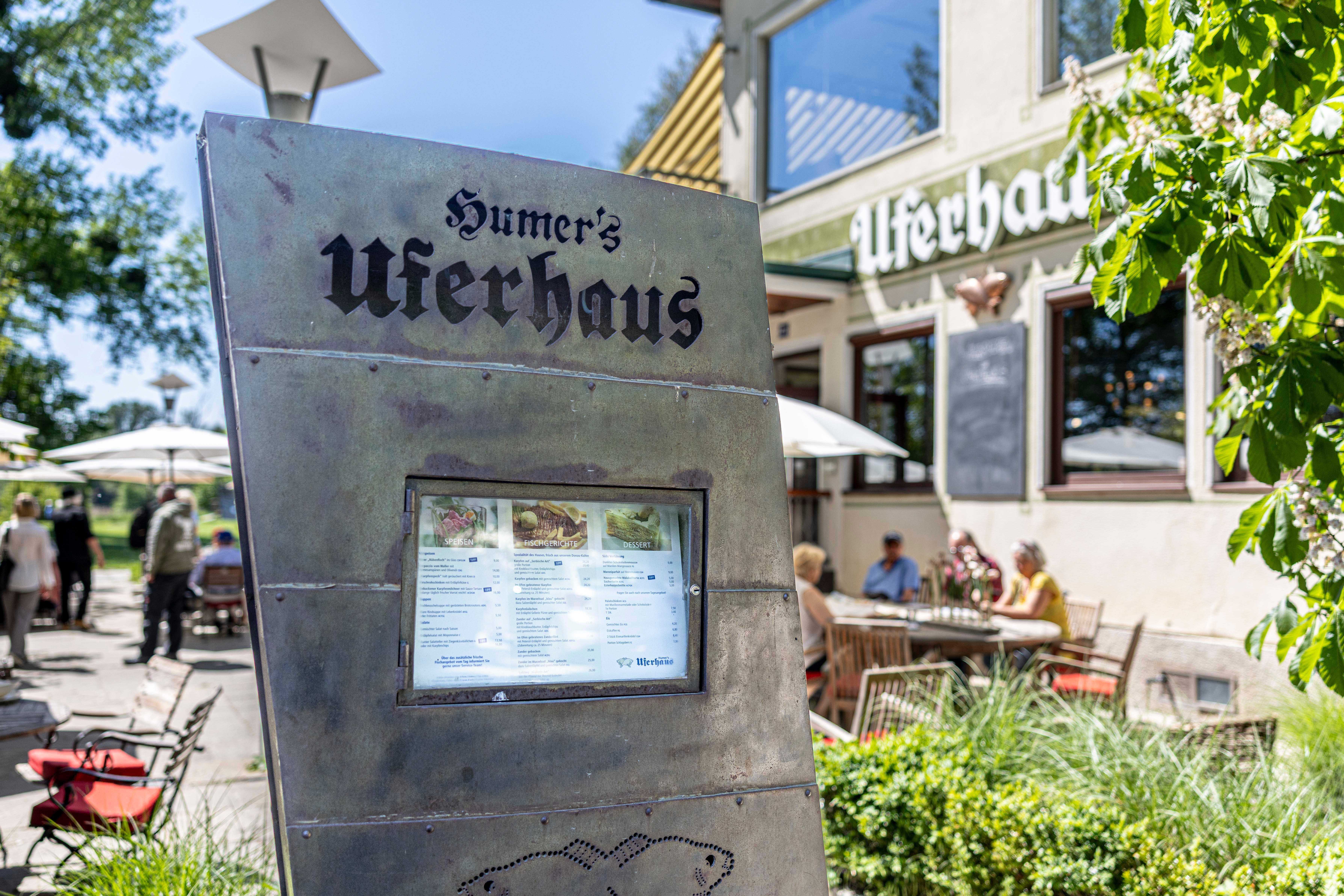 Außenansicht von Humers Uferhaus mit Speisekarte im Vordergrund und Gästen im Hintergrund.