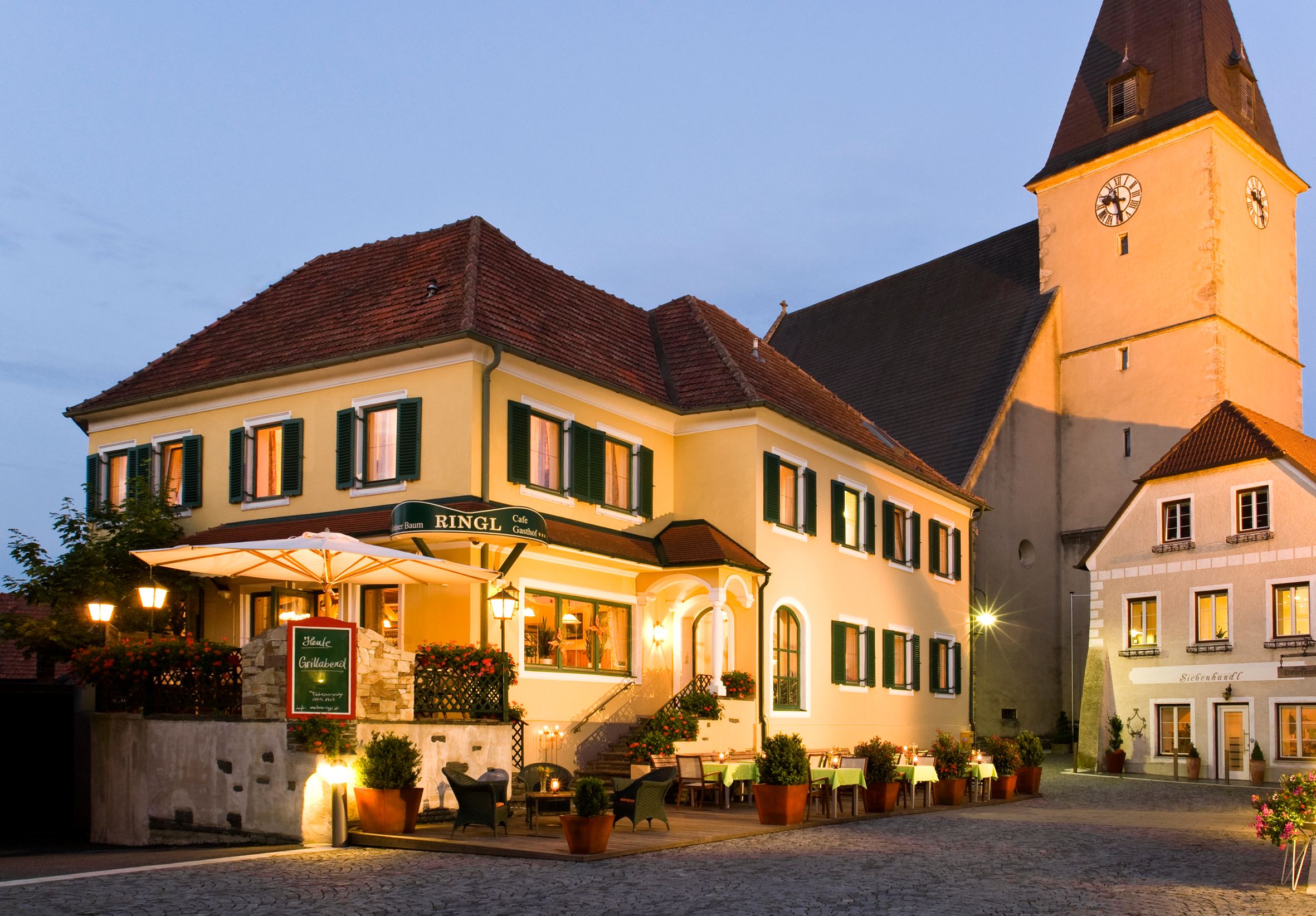 Ein traditionelles Gasthaus mit gelber Fassade und grünen Fensterläden, beleuchtet in der Abenddämmerung, neben einer Kirche.