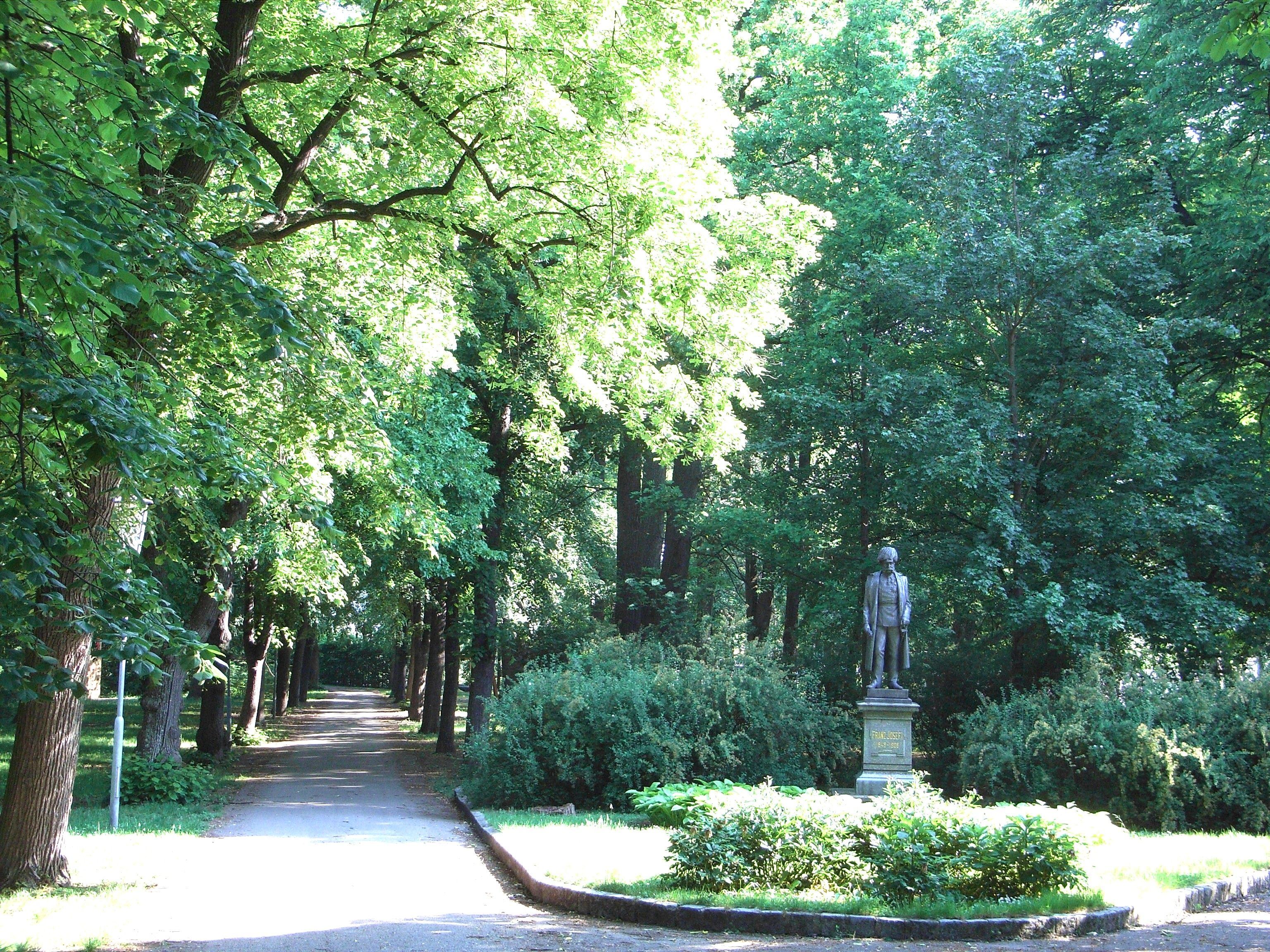 Ein sonniger Parkweg mit einer Statue auf der rechten Seite, umgeben von üppigem Grün.