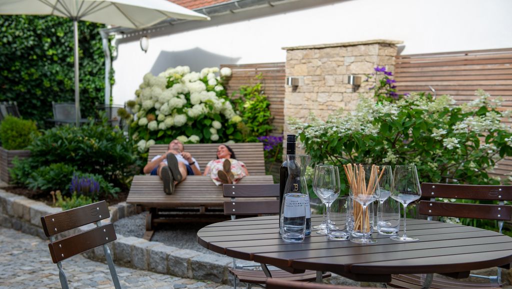 Ein Tisch mit Weingläsern und Flasche, im Hintergrund zwei Personen auf einer Liege im Garten.