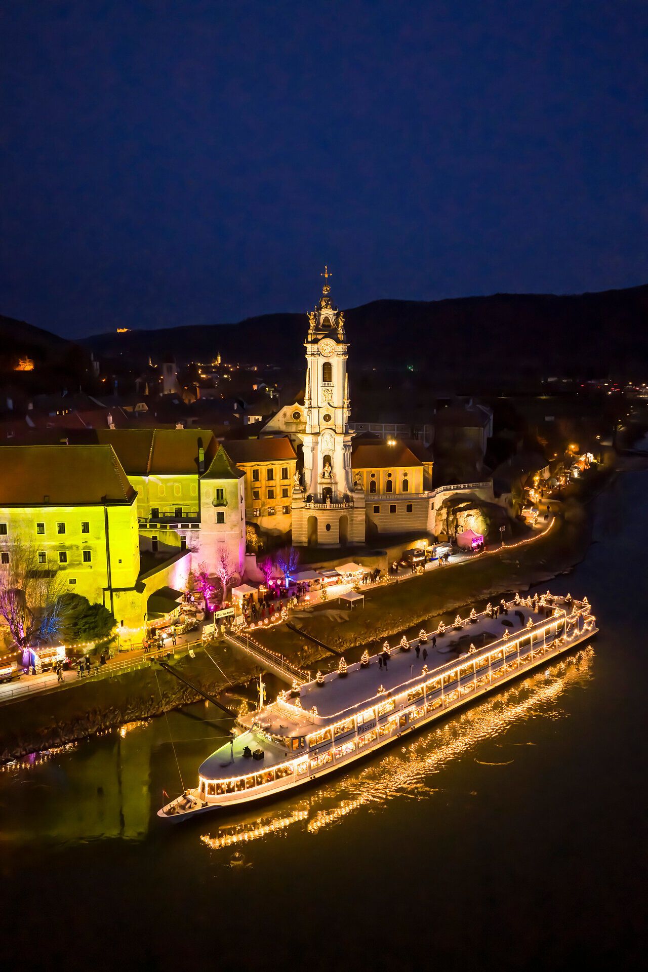 Nächtlicher Blick auf das festlich beleuchtete Dürnstein an der Donau, mit reflektierenden Lichtern, ruhiger Flusslandschaft und historischer Kirche im Advent.