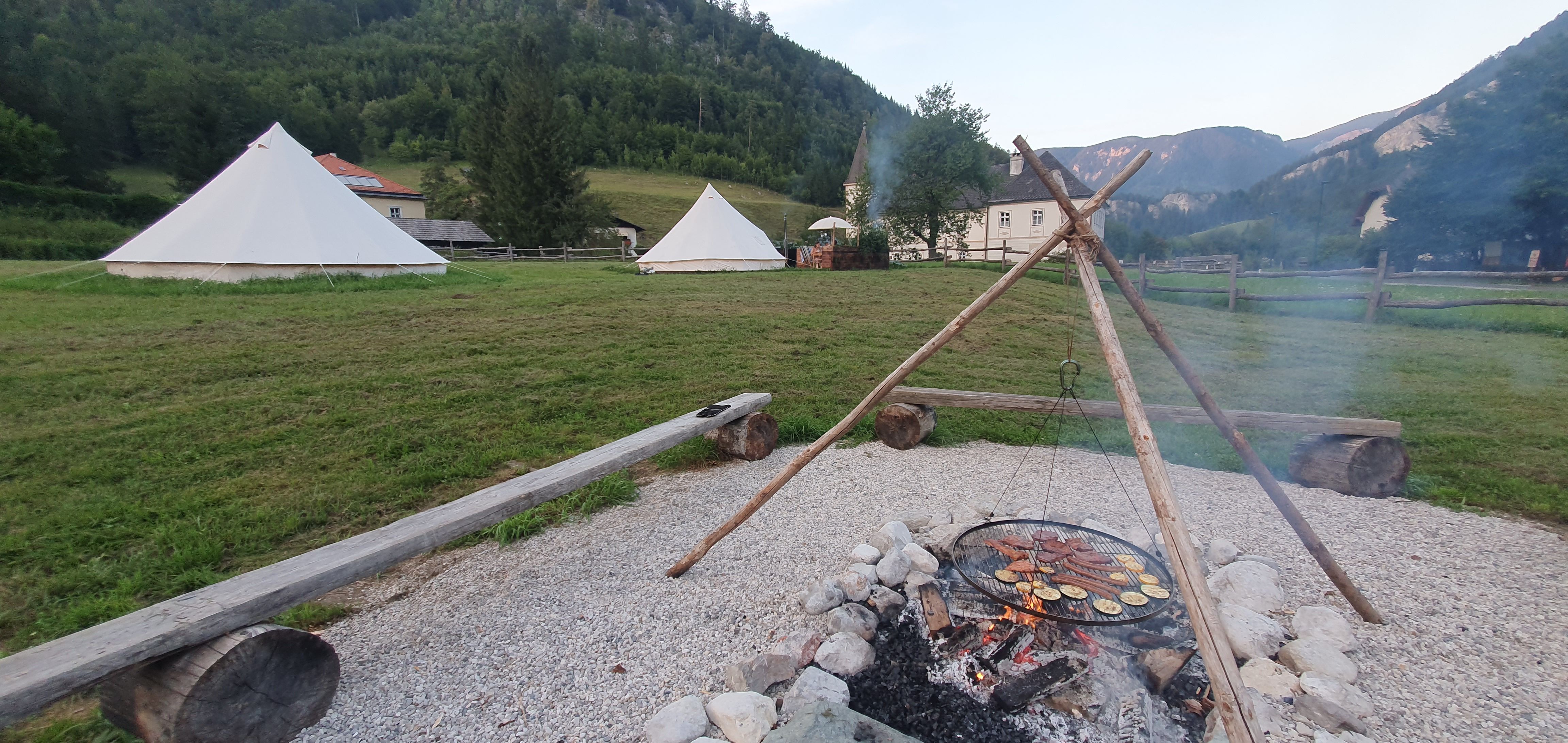Lagerfeuer mit Grill über Feuerstelle, Zelte im Hintergrund, umgeben von Bergen und Wiesen.
