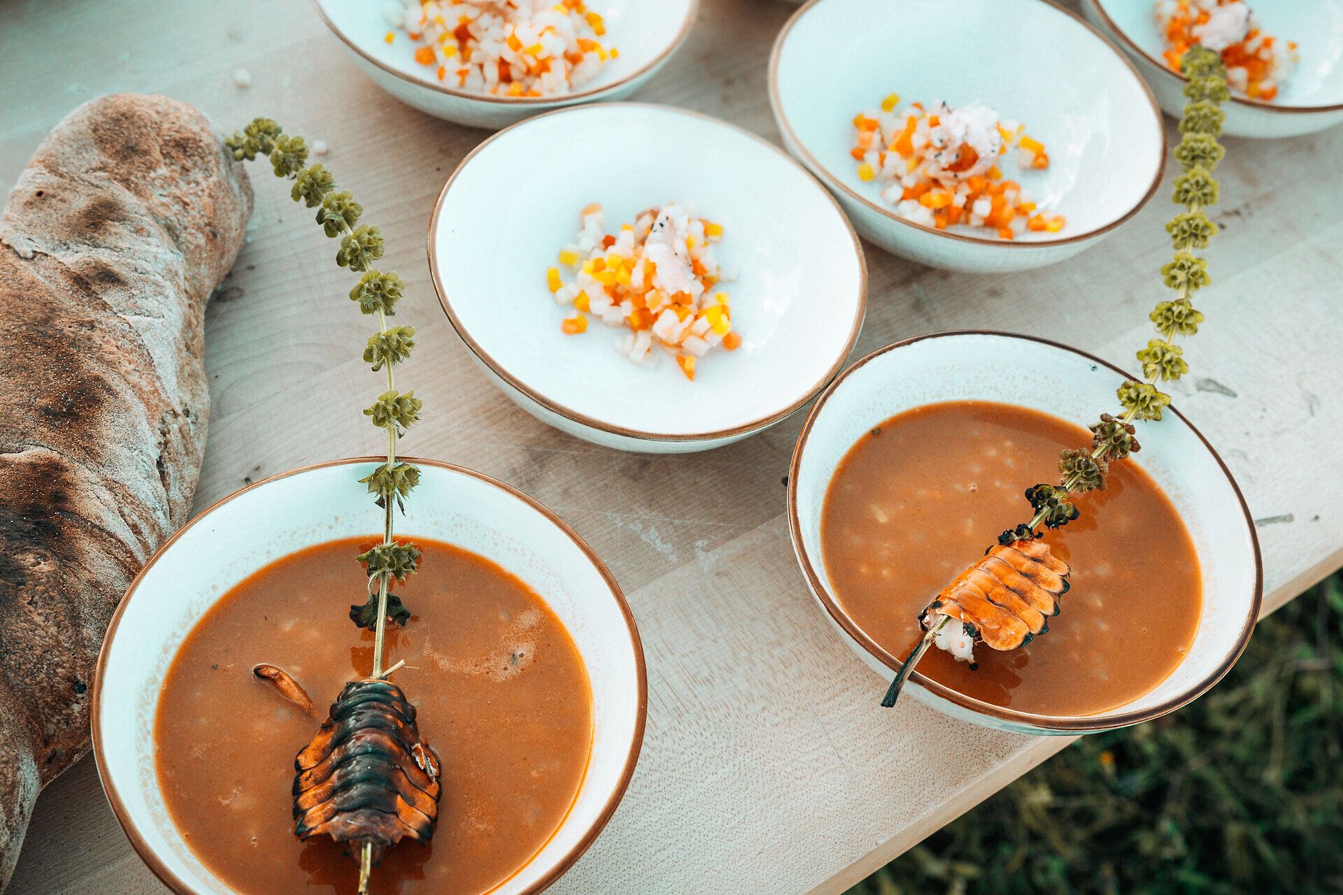 Ein rustikales Festmahl erwartet die Gäste, mit einer verführerischen Suppe, die mit frischen Kräutern und einer besonderen Zutat verfeinert ist. Die kunstvoll angerichteten Schalen laden dazu ein, die Aromen der Region zu entdecken und die kulinarische Vielfalt zu genießen. Ein Stück Brot, das die Tradition der ländlichen Küche verkörpert, rundet das Erlebnis ab.